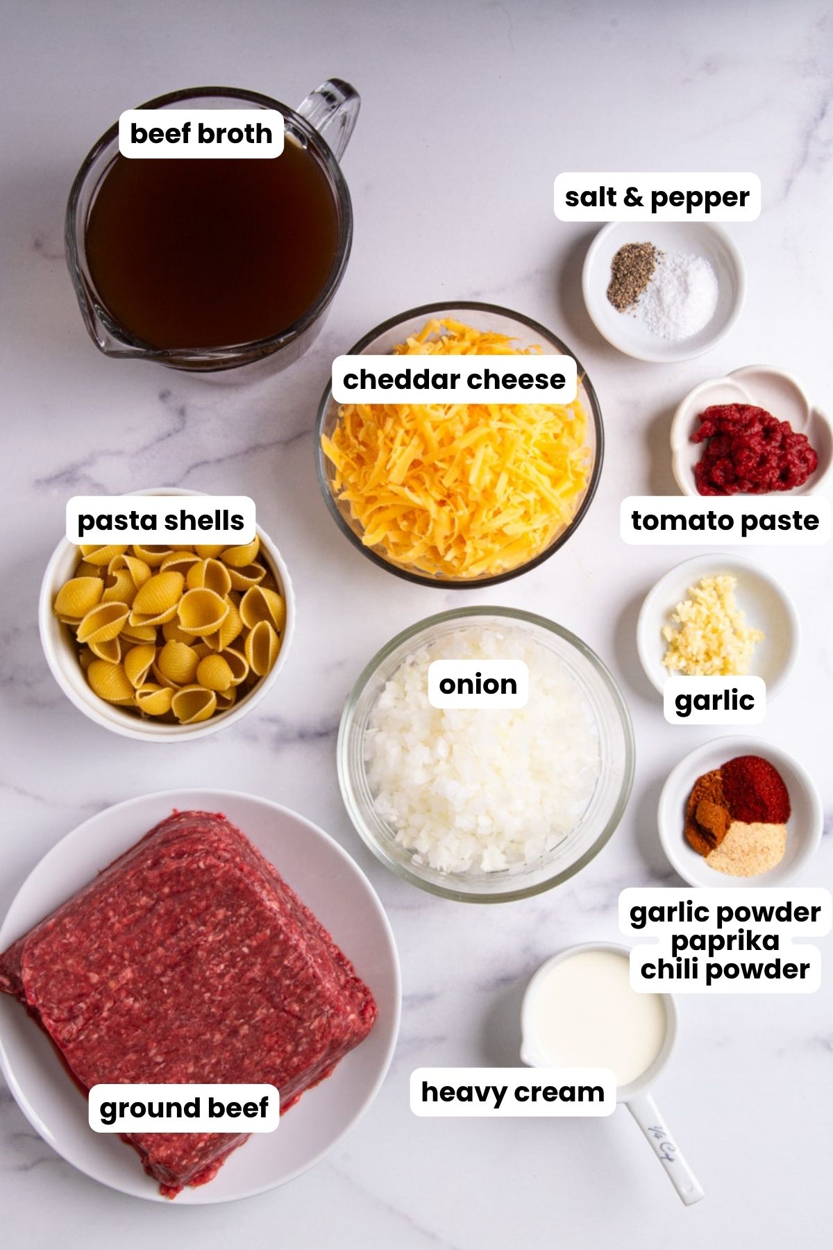 Overhead flat lay of homemade hamburger helper ingredients on a marble surface: beef broth, shredded cheddar cheese, pasta shells, ground beef, diced onion, heavy cream, tomato paste, minced garlic, garlic powder, paprika, chili powder, and salt and pepper, each labeled with text overlays.