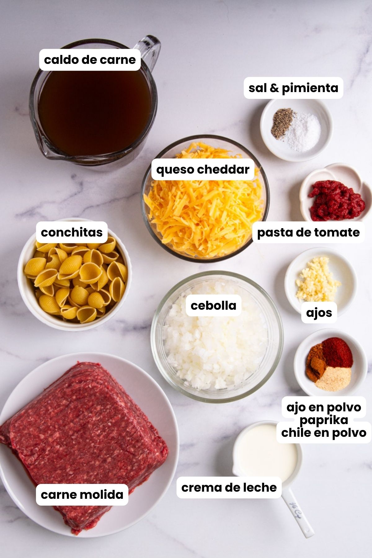 Overhead flat lay of homemade hamburger helper ingredients on a marble surface: beef broth, shredded cheddar cheese, pasta shells, ground beef, diced onion, heavy cream, tomato paste, minced garlic, garlic powder, paprika, chili powder, and salt and pepper, each labeled with Spanish text overlays.